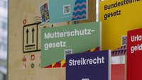 Das Bild zeigt eine Detailaufnahme einer Ausstellungseinheit der Vielfaltsausstellung, die verschiedene Plakate mit gewerkschaftlichen Themen darstellt. Zu sehen sind Plakate mit den Titeln "Mutterschutzgesetz", "Streikrecht", "Bundesteilhabegesetz" und "Urlaubsgeld", die jeweils mit einem QR-Code versehen sind, der zu weiterführenden Informationen führt. Die Plakate sind in lebhaften Farben gestaltet und stehen auf einem roten Untergrund, der den Slogan "Für Vielfalt erkämpft" trägt. Diese Präsentation hebt die Bedeutung und Errungenschaften der Gewerkschaften in verschiedenen sozialen und arbeitsrechtlichen Bereichen hervor.