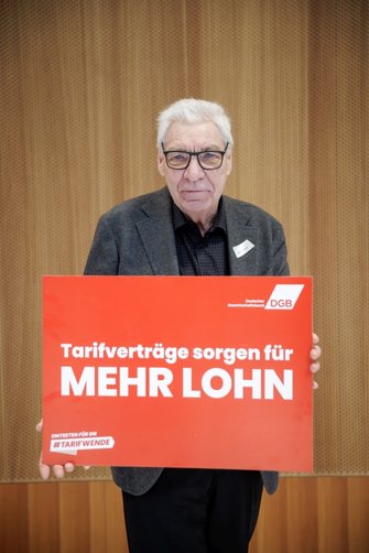 Foto von Michael Sommer, ehemaliger DGB-Vorsitzender, der ein DGB-Plakat in den Händen hält mit der Aufschrift "Tarifverträge sorgen für MEHR LOHN". 