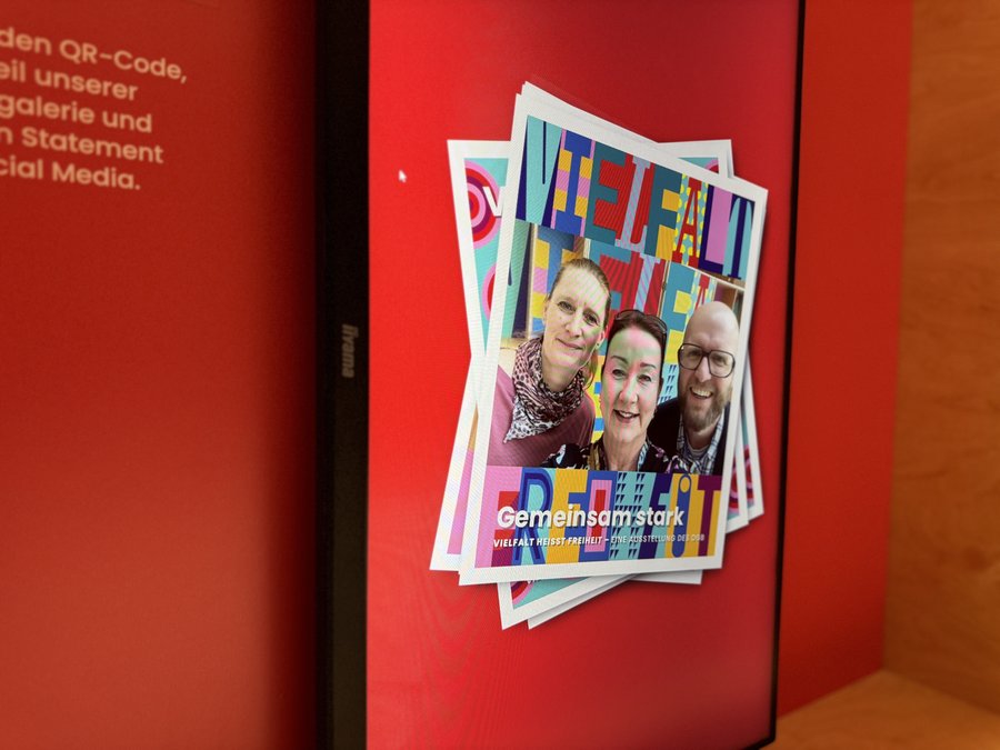 Das Bild zeigt einen Bildschirm, auf dem mehrere übereinanderliegende Selfies abgebildet sind. Die oberste Karte trägt die Wörter "Vielfalt Freiheit" in bunten, stilisierten Buchstaben. Der Hintergrund des Bildschirms ist rot, und links neben dem Bildschirm befindet sich Text, der Informationen über die Nutzung eines QR-Codes für den Zugang zu einer Online-Galerie und die Möglichkeit zur Teilnahme auf Social Media bietet. Die Gestaltung ist lebendig und auffällig, um das Thema Vielfalt und Gemeinschaft zu betonen. Der Text "Gemeinsam stark" ist ebenfalls sichtbar und unterstreicht die Botschaft der Einheit und Stärke.
