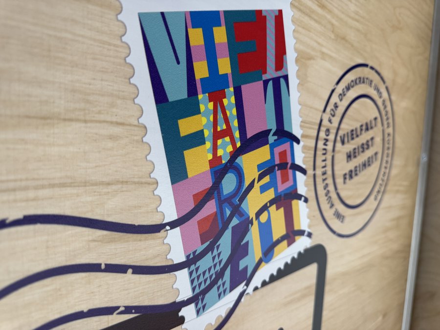 Das Bild zeigt eine Nahaufnahme eines Holzpaneels mit einem bunten Aufkleber, der die Worte "Vielfalt Freiheit" in großen, stilisierten Buchstaben darstellt. Der Aufkleber ist im Design einer Briefmarke gehalten und zeigt lebhafte Farben wie Blau, Rot, Gelb und Violett. Neben dem Aufkleber ist ein runder Stempel zu sehen, auf dem "Vielfalt heißt Freiheit – Eine Ausstellung für Demokratie gegen Ausgrenzung" steht. Das Design ist auffällig und farbenfroh, um das Thema Vielfalt und Freiheit zu betonen.